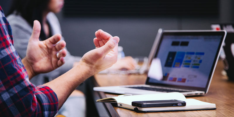 Man gesturing in front of a macbook and a notebad and iphone on the desk