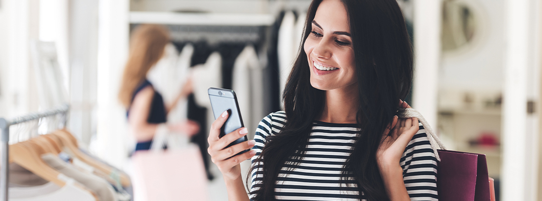 woman smiling at mobile phone screen holding shopping bag