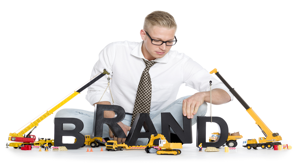 Build up a brand concept: Smiling businessman building the word brand along with construction machines, isolated on white background.