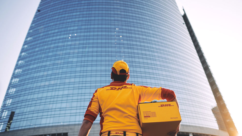 DHL delivery man holding parcel and looking up to glass building