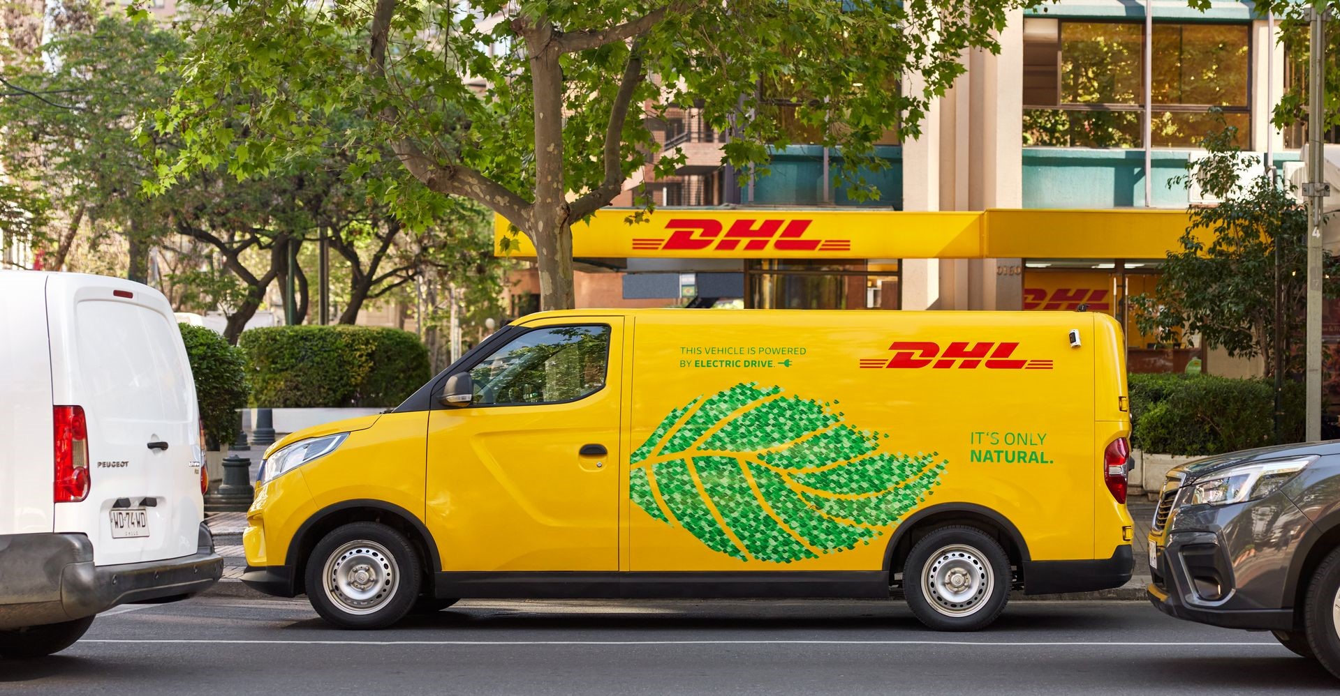 An electric DHL van is standing outside a DHL service point.